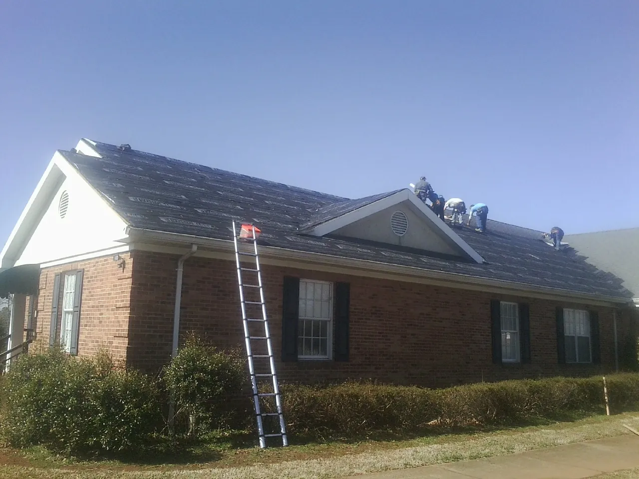 Crew tearing shingles off a brick ranch roof with a ladder set — Metal Roof Repair work by our crew in Conrad
