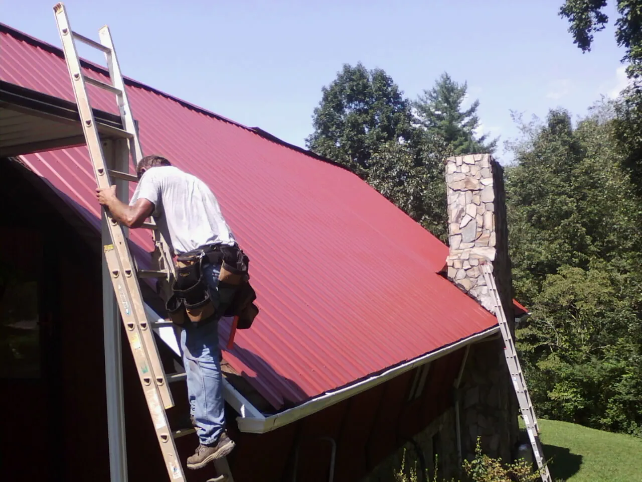 Roofer on a ladder beside a red metal roof and stone chimney — Soffit & Fascia Repair by Roofing Brothers in Conrad